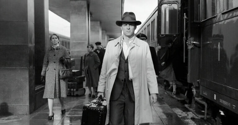 Black and white photo of Andrew Scott in a trench coat and fedora at a train station.