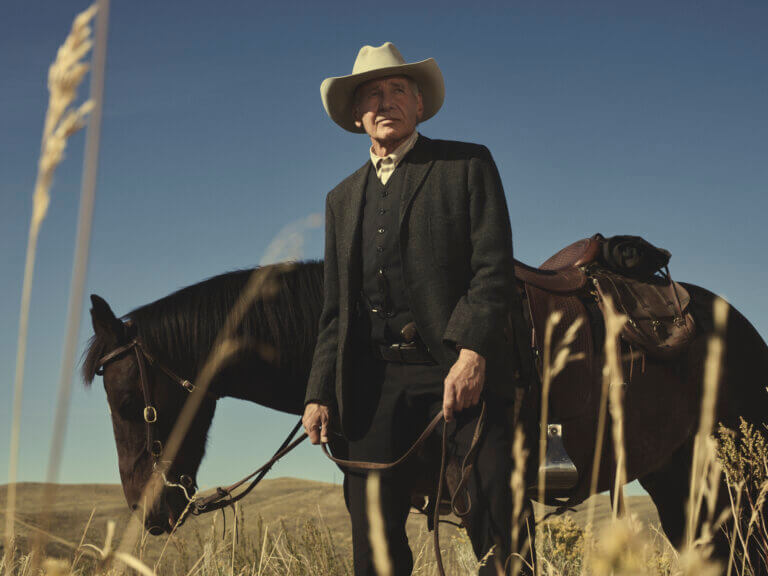 Harrison Ford in black cowboy gear outside with a black horse.