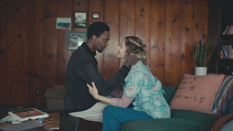 Two actors portraying a scene as an interracial couple embracing in a room with wood walls.