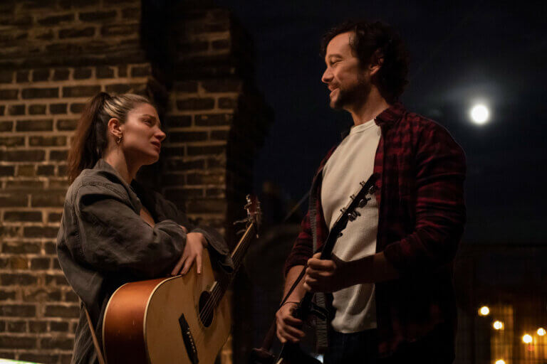 Eve Hewson and Joseph Gordon-Levitt inside talking and holding guitars.