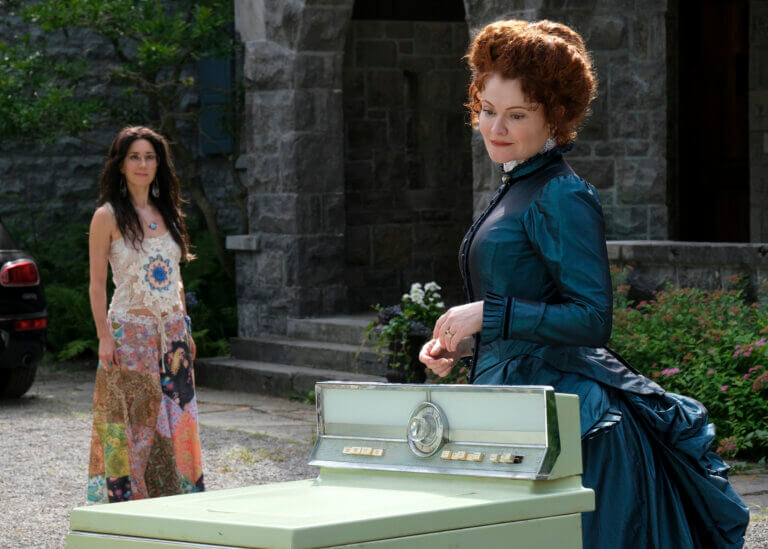 Actors Sheila Carrasco as Flower and Rebecca Wisocky by a white piano in a garden on CBS' "Ghosts."