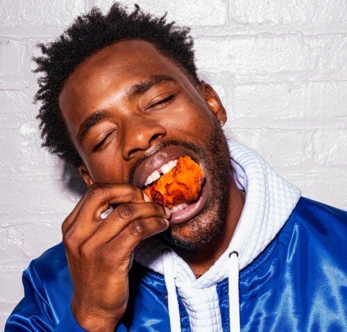 Actor John Adewumi eatching a chicken wing wearing a blue jacket over a hoodie.