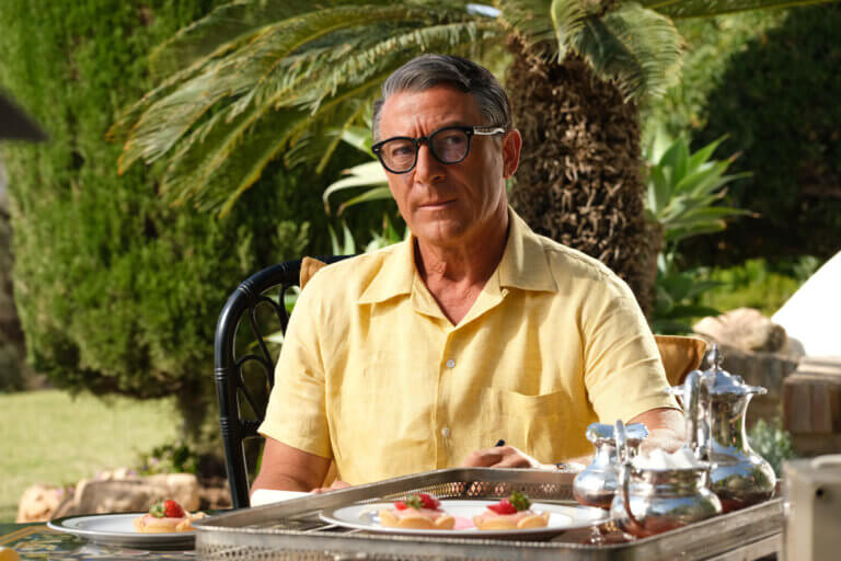 Jason Isaacs as Cary Grant in a yellow shirt sitting outside.