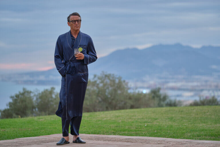 Jason Isaacs as Cary Grant in a bathrobe having a drink outside.