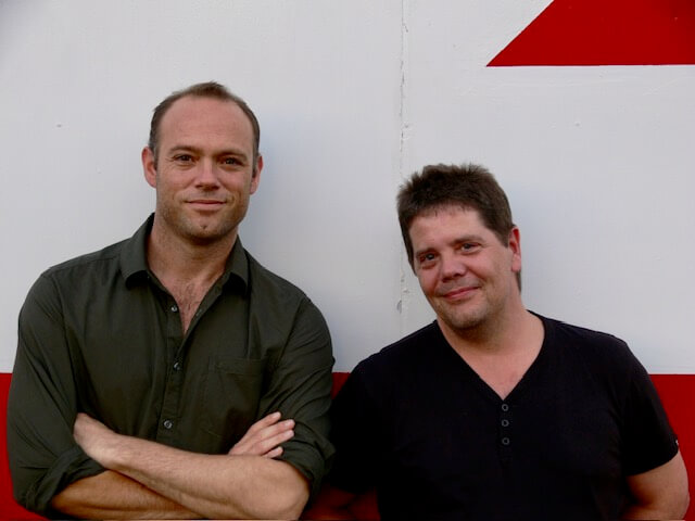 Tom McSweeney & Dave Newman standling against a white and red wall smiling.