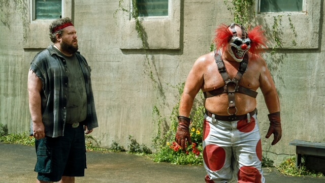 Mike Mitchell in a cutoff flannel shirt outside with a scary muscular clown.