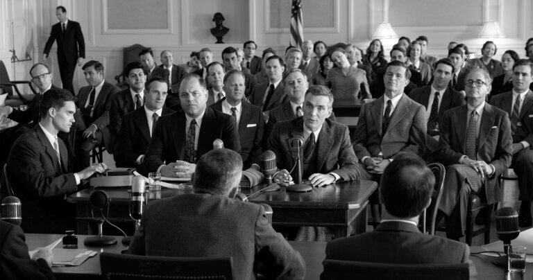 Black and white still from the movie Oppenheimer. A press room of curious reporters and a man on the stand.