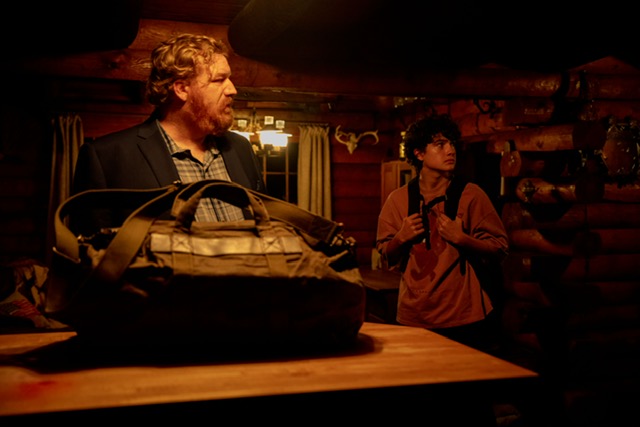 Happy Anderson looking surprised in a cabin standing over a table with a duffel bag on it.