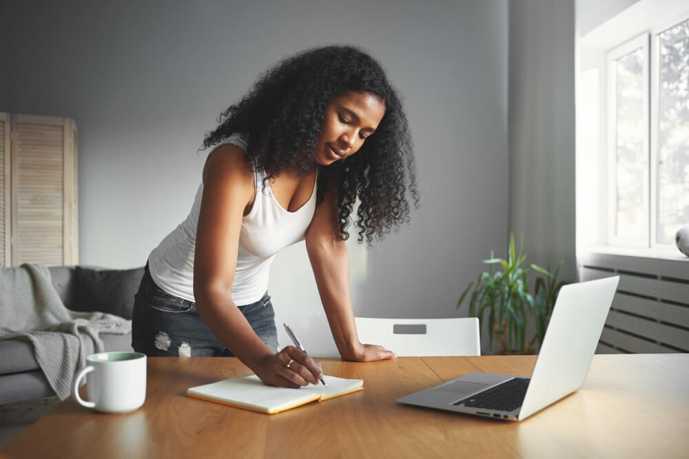 African American actress in a tank top making plans on her laptop and writing it down in her notes at home.