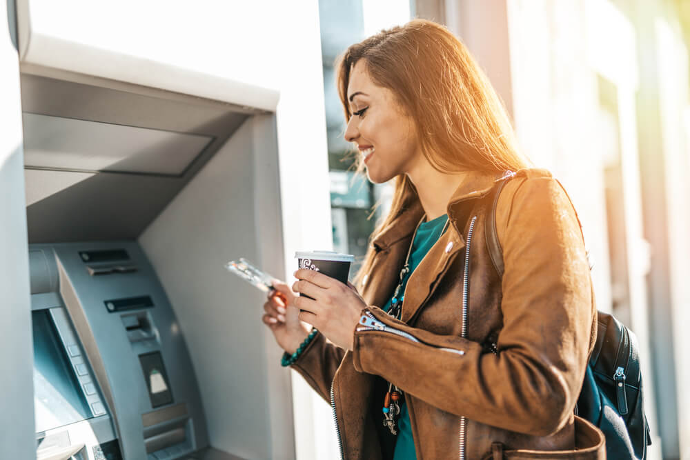 Happy woman, coffee in hand, withdrawing money from an ATM outside.