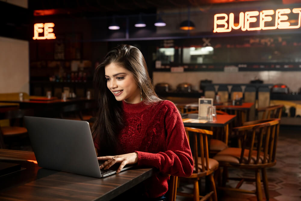 An Indian woman in a red shirt at a restaurant on her laptop.