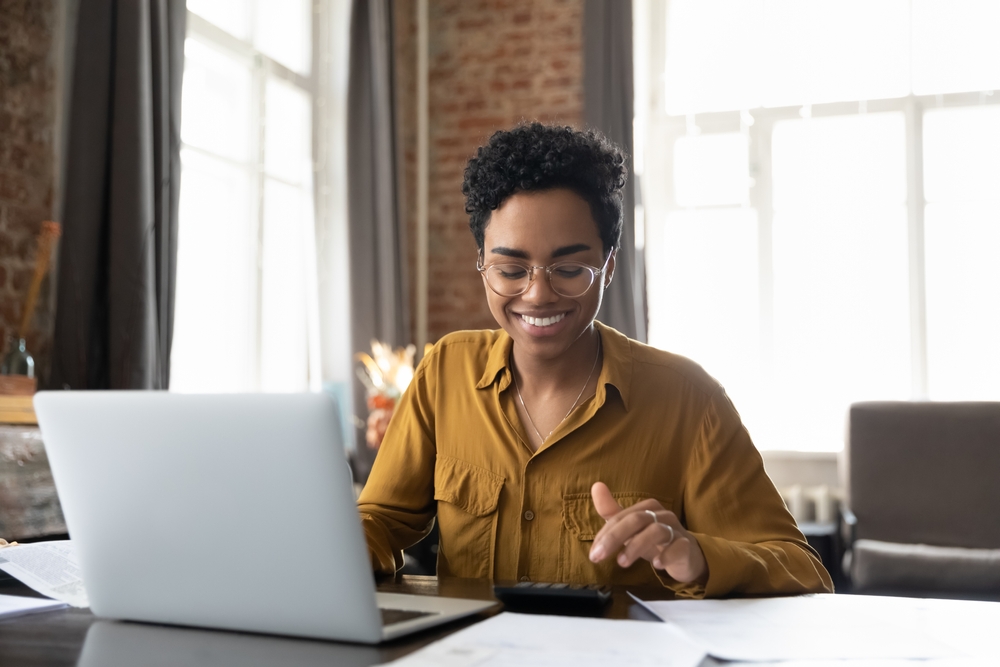 Happy African American woman crunching numbers on her laptop inside.