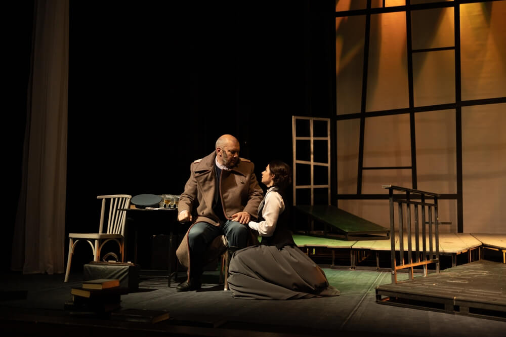 Two actors in old clothes performing in a play surrounded by chairs and wood pallets.