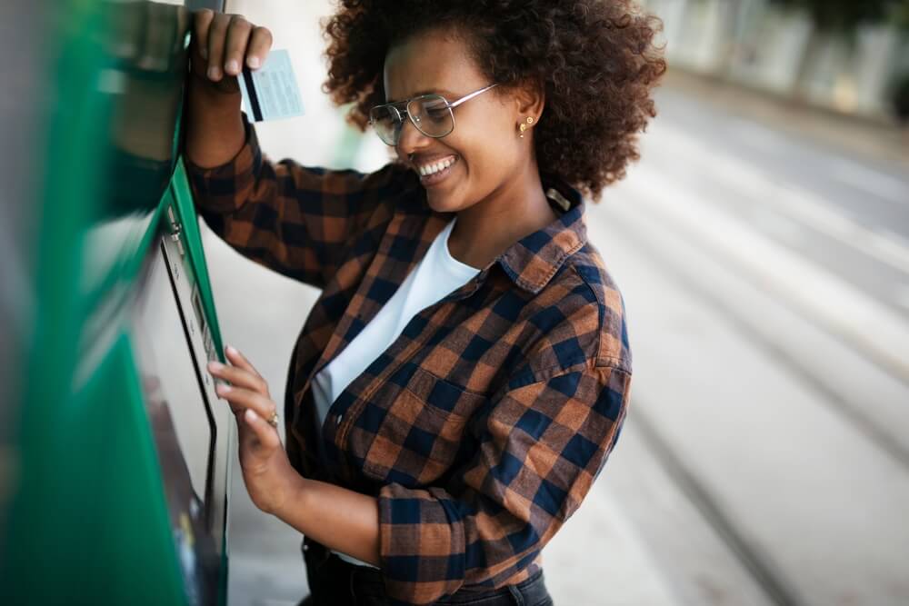 African American woman in a flannel shirt smiling while using a green ATM machine.