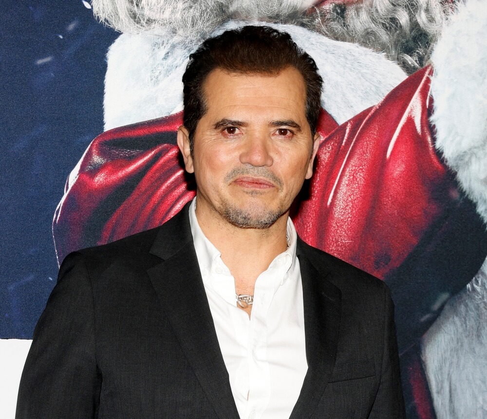 Actor John Leguizamo in a suit standing in front of a backdrop smiling.