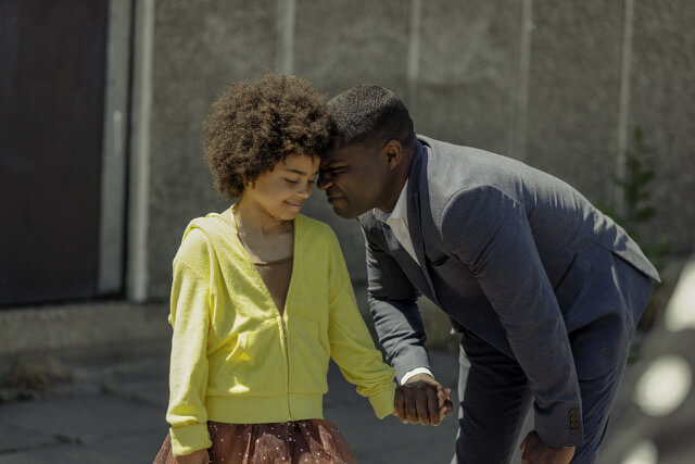 David Oyelowo in a suit smiles and holds his daughter's hand.