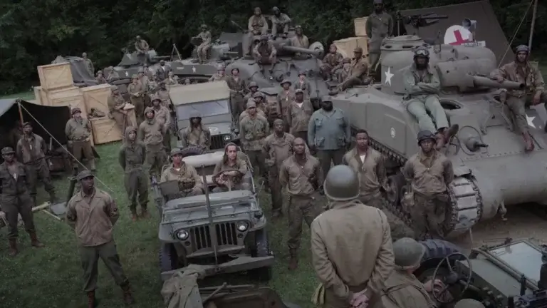 A group of military men in army fatigues at a base  surrounded by army cars and a tank awaiting instructions.